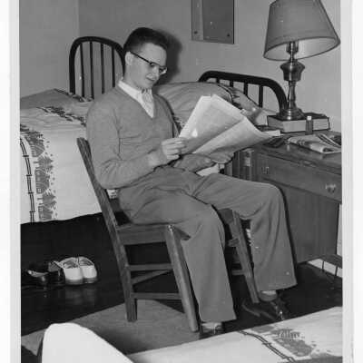 Student of Quirk Hall reading newspaper in dormitory room.