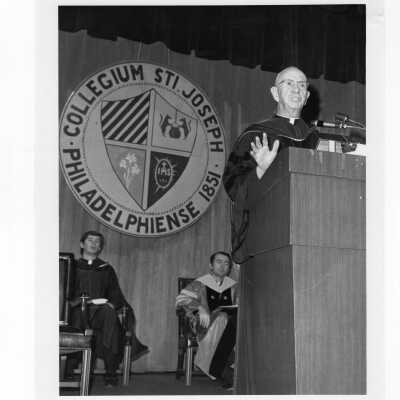 Dennis Comey, S.J., addressing convocation, December 17, 1975, with Edward Denion,S.J., and Thomas F. Gleeson, S.J. in background.