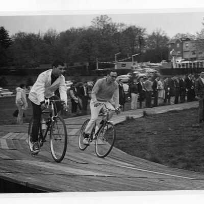 Spectators watching two students in bicycle race on wooden track near Field House during Junior Week activities.