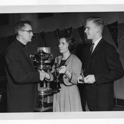 Student from Villiger Debating Society and woman presenting trophies to Jesuit (?) with TV Guide trophy in background.