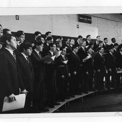 Students in Glee Club wearing coat and tie and holding sheet music practicing.