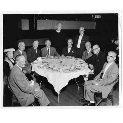 View of table with SJC graduates in early 20th century with J. Joseph Bluett, S.J., at alumni communion breakfast, October 28, 1956. Includes, E. Dougherty, H. Flanigan, N. Considine, L. Frank, L. Heine, T. Welsh, F. Hayes.