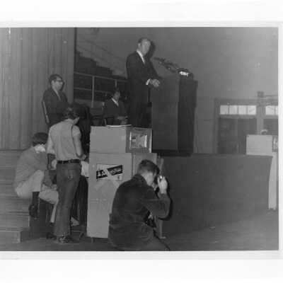 Vice President Hubert Humphrey speaking from podium in Field House during his visit to SJC campus, January 27, 1970, with Dan DeLucca and several students behind and to side of stage.