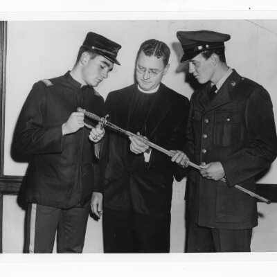 Edward G. Jacklin, S.J. with Tom Duffy in SJC cadet corps uniform and Bill Berry in ROTC uniform during centennial celebration event held October 21-28, 1951. The three individuals are looking at sword and scabbard (on loan from Breen family).