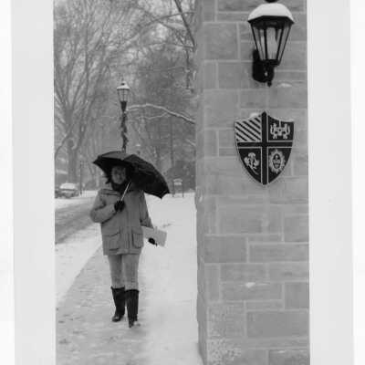 Woman student walking down Lapsley Lane in snow near entrance pillar with seal.