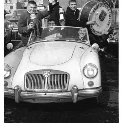 Students, including John Lehman and Al Tegler, driving in convertible automobile during booster club pep rally. Lehman is driving and Tegler is holding Air Force ROTC drum.