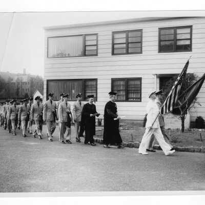 ROTC graduating students processing in uniform in front of cardboard college to 1955 graduation/commencement ceremony. Two black and white prints.