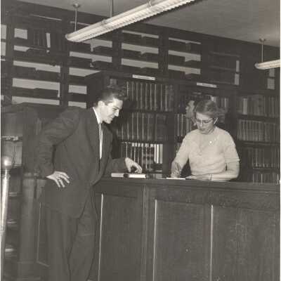 Interior view of library in Barbelin building with student checking out book at circulation desk and talking to librarian.