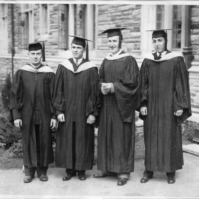 View of four graduates in dressed in cap and gown either before or after 1928 graduation/commencement ceremony.