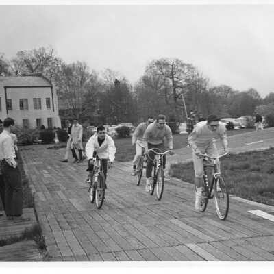 Students Steve Matarazzo, Bill McCullough, Bill Hawes, and team captain Dan Dwyer in bicycle race on wooden track near Field House during Junior Week activities, Spring 1966. Part of a 24 hour overnight event.