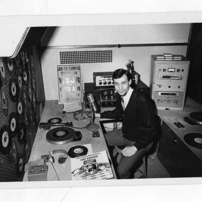 Student member of Radio Club sitting at Station WSJR broadcasting and playing records. Records are hanging on hooks on wall pegboard.