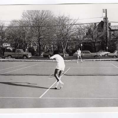 Two members of men's tennis team practicing on court with cars parked on street and neighborhood in background.