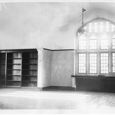 Interior view an empty room in Barbelin with built-in bookcases that are empty, a radiator, arched leaded windows, and chandelier.