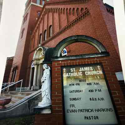 Colorized exterior photo of the St James Catholic Church entrance. The mass schedule and name of the pastor or listed on the signboard outside.