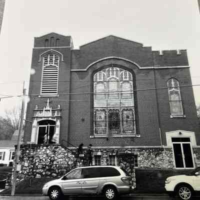 Black and white exterior photo of a brick and cobbled stone church with tall stained glass windows, a squat turret above the entrance door, and parapet style roofing accents.