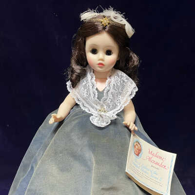 The image features a Madame Alexander doll dressed in a grey dress. The doll has long, dark curls and wears a headband embellished with feathers. Her attire consists of intricate lace detail around the neckline. The doll resembles Angelica Van Buren, daughter-in-law to president Van Buren.