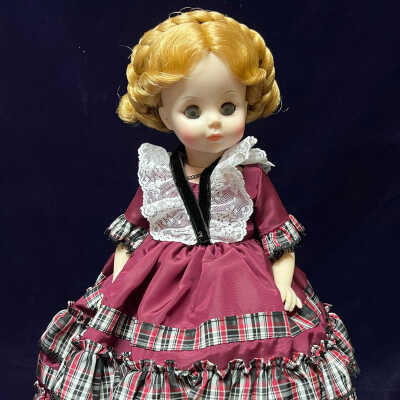 The image features a porcelain doll of Betty Taylor Bliss, the daughter of Zachary Taylor. The doll has curly blonde hair styled in an intricate updo. The doll is dressed in an elaborate burgundy gown adorned with layers of ruffled plaid fabric in red, black, and white. Additional details include a delicate white lace collar and black velvet trim accentuating the dress. The doll's facial features are delicately painted, with large, bright eyes and subtle blush on its cheeks.