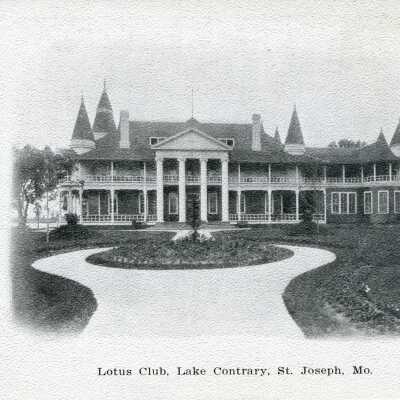 Lotus Club, Lake Contrary, c. 1910: Black-and-white photo postcard depicting the front view of the Lotus Club at Lake Contrary, showcasing a grand two-story white building with tall pillars, a wraparound porch, balcony, steeple turrets, and a circular lane with a central flower garden.