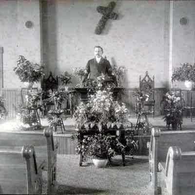 Man standing on church platform in front of pews; 15 floral arrangements