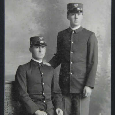 ¾ figure Portrait 2 males; 1 standing and 1 sitting, both in uniform wearing hats