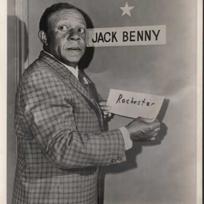 Photo - Jack Benny, B & W. in front of dressing room, holding a sign that says "Rochester" .