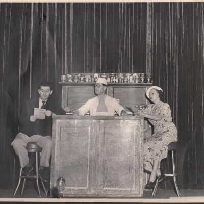 Photo - Resturant type scene B & W-Comic behind bar, male and female seated on either end.