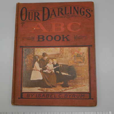 Hardback book with red cover, multicolored plaque on front depicting a woman in a long, dark skirt reading to children