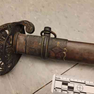 The image depicts a close-up view of a decorative sword hilt and sheath featuring intricate engravings and patterns, with a detailed guard showcasing swirling, ornate designs. A measuring scale lies beside the sword on a patterned, light-colored floor, indicating the sword's dimensions. The metal of the sword appears aged with a patina, adding to its historical allure.
