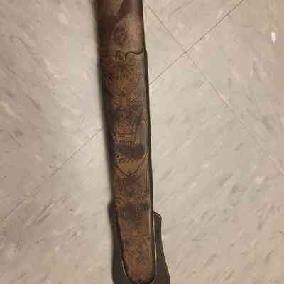An intricately engraved sword sheath rests on a speckled tile floor, showcasing decorative patterns.