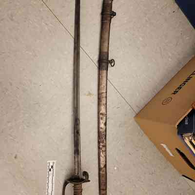 The image showcases a historical sword, from the Civil War era, attributed to Colonel William Mungen of the 57th Ohio Volunteer Infantry (OVI). The sword lies next to its scabbard. The handle of the sword is wrapped, accompanied by a guard that features intricate metalwork. The scabbard, situated to the right of the sword, is adorned with ornate details and includes metal fittings and rings for attachment.