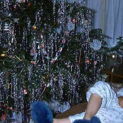31. KJH sitting by decorated Christmas tree with blue slippers: Zylstra House - Dec. 1960