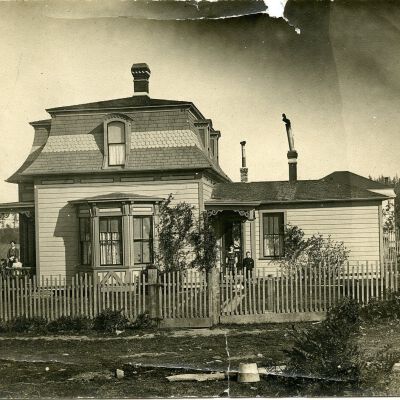 2019.042.307: Photo of a two-story building with mansard roof in different colors, central chimney; other chimneys on the one-story addition on the right. Dormer windows. Bay window in front. Portico over front door with decorative trim and a woman and two children on the porch. In front of the addition is a small portico over a door, decorative trim and two children. Picket fence. Laundry drying on the right. On the back: O. S. Van Olinda, Coupeville, Wash. (H. B. Lovejoy, builder) Captain Howard Lovejoy's home, before we bought it. Now, 1974, on Historical preservation list - Ida May VanOlinda and Marjorie front porch, Myrtle VanOlinda and Eugene on other about 1907.