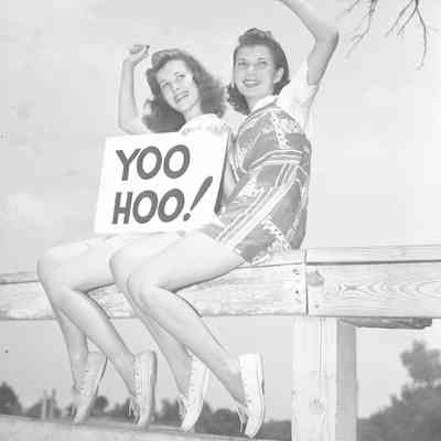 Two girls sitting on fence holding a "YOO HOO" sign