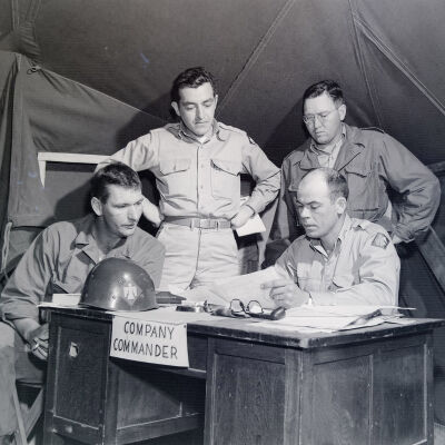 Soldiers gathered around the Company Commander at his desk in a tent