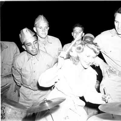 Woman playing drums with soldiers looking over her shoulder