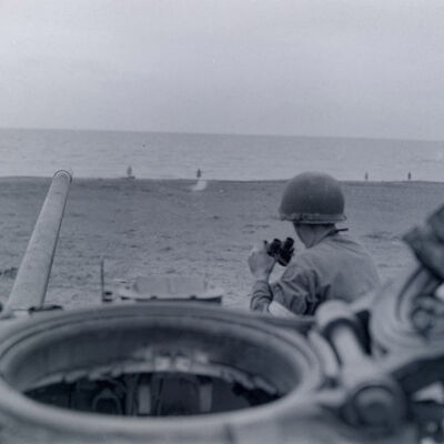 Soldier with binoculars sitting on a tank overlooking a beach area