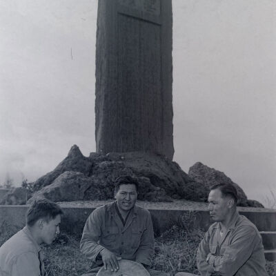 Three Native American soldiers with a drum sit in front a large stone obelisk