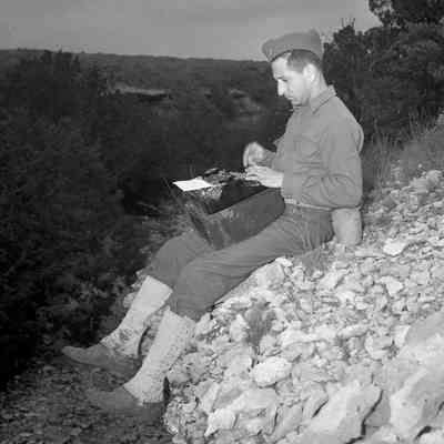 Soldier using a portable typewriter while sitting on a rock ledge