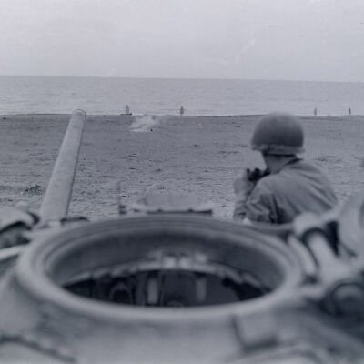Soldier with binoculars sitting on a tank overlooking a beach area