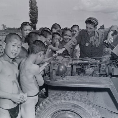 Soldiers looking under the hood of a jeep with a large group of Japanese kids