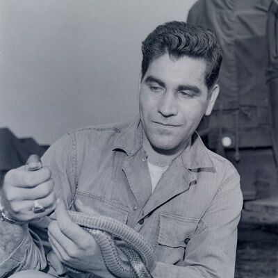 Soldier posing with a snake with a tent camp in background