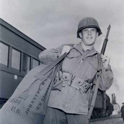 Soldier holding duffel bag and weapon with a train car in the background