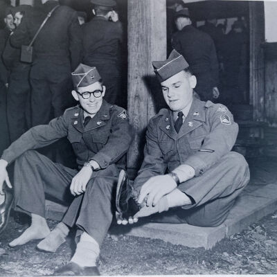 Two soldiers in Class A uniforms sit and take their shoes off