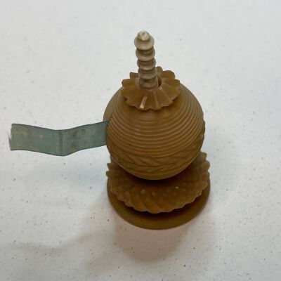 #4: Bone and Tagua nut retractable tape measure. End  of teal tape is missing, now starts at number three. Possibly a top finial is missing as there is a bone spiral pole extending form the center of the ball-shaped tape measure. The nut is carved with a wreath of leaves along the bottom of the ball-shape and coils surrounding the shape to the top where a flower-like collar sits. Tehre is a slender slot on the side for the tape to be pulled out.
