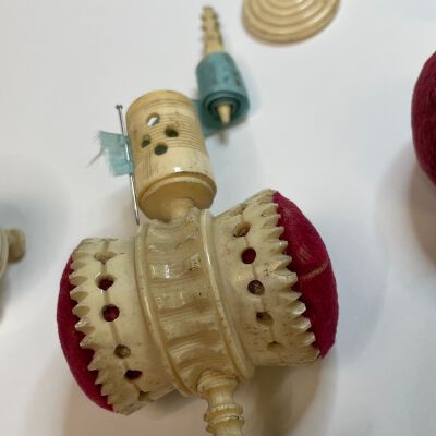 #3: Carved bone pedastal with double barrel red velvet pin cushions extending at the center sides in a horizontal bar. The top has four circles carved into it where a staff is inserted with a teal hand-numbered measuring tape. the top is a cap with a hole where the finial of the tape measure winder protrudes.