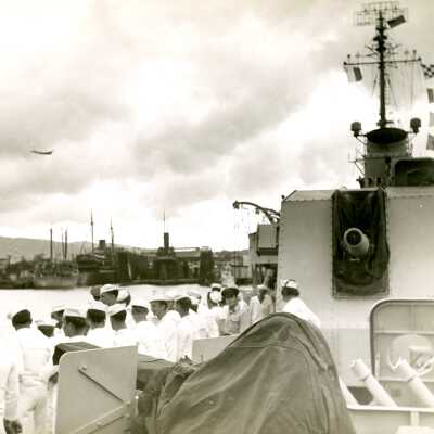 2019.06.48-5: Men in white sailors' uniforms getting ready to disembark at Pearl Harbor. 5'-gun turret is facing the camera and aircraft is flying in background.