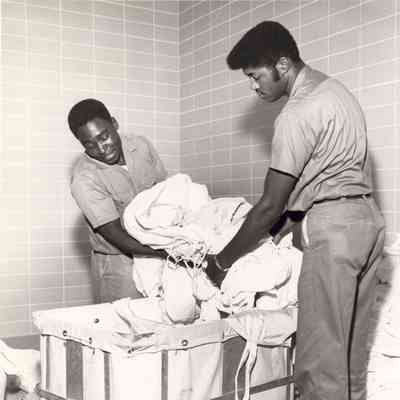 Two men in uniform are handling a large pile of laundry, smiling and working together efficiently.