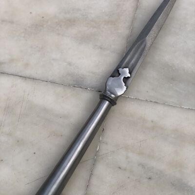 The image displays a spearhead featuring a sharp, elongated metal blade with a distinctive, jagged cut-out near the base, attached to a smooth, metallic shaft that transitions into a textured wooden handle, all resting against a mottled gray tiled floor.