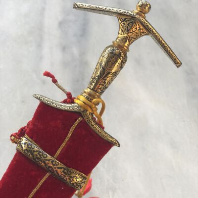 Ornate ceremonial dagger with detailed gold designs emerges from a red velvet sheath adorned with gold trim