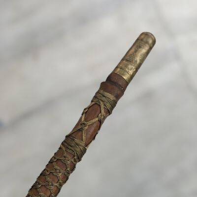 The image displays a close-up of a traditional artifact, potentially a walking stick or ceremonial staff, featuring intricate craftsmanship. Its handle is elaborately adorned with metal and leather, wrapped in a distinctive, tightly bound pattern of metal wire that forms crisscrossing and looping designs over the length of the artifact. The handle's top, slightly wider than the stem, shines with a golden hue and is engraved with detailed motifs that suggest cultural or ceremonial significance, possibly originating from an indigenous or ancient community. The blurred background helps to isolate and highlight the craftsmanship and historical aesthetics of the staff.